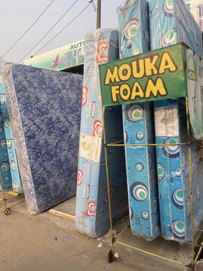 This image captures an outdoor commercial scene in the city of Isasi, Nigeria, specifically an open-air mattress retail display. The foreground and midground are dominated by numerous new mattresses, most wrapped in clear protective plastic.
On the left, a large, dark blue mattress with a quilted, intricate floral pattern stands upright. To its right, a series of light blue mattresses, also plastic-wrapped, are arranged, featuring a distinctive pattern of concentric circles in white, light blue, and dark blue, alongside red and white floral motifs. Many of these mattresses prominently display "MOUKA FOAM" branding on their packaging.
A central green sign with yellow block letters spelling "MOUKA FOAM" is mounted on a yellow metal display rack, which supports several vertically stacked mattresses. Additional yellow metal racks, appearing to be constructed from rebar and fitted with small wheels, are positioned around the display, holding more mattresses.
In the background, a large white sign with green lettering is partially visible, indicating an "AUTHORISED DEALER" and displaying a telephone number (0915...). This sign also features graphic depictions of individuals carrying mattresses. Overhead power lines are visible against a pale, clear sky. The ground surface is composed of worn concrete or asphalt, with scattered small pieces of debris. No individuals are present in the immediate vicinity of the display.