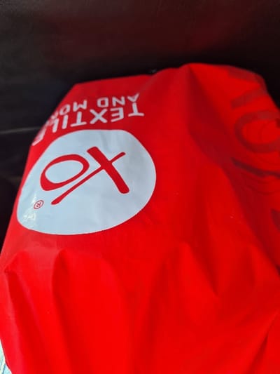 The image displays a red shopping bag, made of a flexible, possibly plastic or fabric material, positioned at an angle within a dimly lit environment. A prominent white circular area on the bag features a stylized red "OX" logo, accompanied by a registered trademark symbol (®). Above this logo, inverted white text reads "TEXTILE AND MO," suggesting the full phrase "TEXTILE AND MORE." The bag exhibits folds and creases across its surface. The background is dark, out of focus, and indistinct, indicating an interior setting, possibly a vehicle interior or a contained space. No individuals, actions, or interactions are visible. The specific location within the city of Ma'raba, Syria, cannot be determined from the visual content of the image.