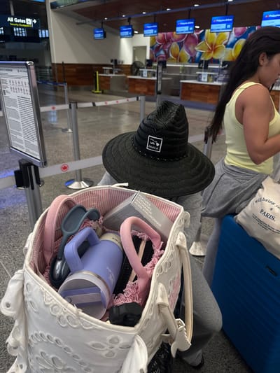 The image was taken at Honolulu Airport, United States of America. It shows a woman wearing a yellow tank top and grey sweatpants seated next to a blue suitcase and a white tote bag. Inside the tote bag, among other things, are a purple Stanley water bottle, an Apple Watch, a grey box of skincare, a pink headband, and a wide-brimmed black hat. The background shows airport signage.