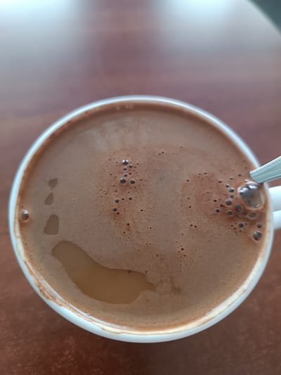 A close-up, overhead shot shows a white ceramic mug filled with hot chocolate sitting on a brown wooden table. The hot chocolate has a foamy surface with small bubbles, and a spoon is partially visible on the right side of the mug, submerged in the drink. The lighting suggests an indoor environment. The image's composition is centered on the mug, with the background slightly blurred. There are no people or animals visible in the image. The overall scene depicts a simple, everyday moment of enjoying a beverage. The provided location of Skidzieĺ, Belarus is not visually evident in the image itself and would require external information to confirm.