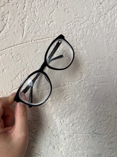 A pair of black-rimmed eyeglasses is being held by a person's hand against a textured, light-colored background. The eyeglasses have clear lenses and are positioned at an angle. The background appears to be a wall with a rough, uneven surface, possibly stucco or plaster. The lighting suggests an indoor setting, possibly during the daytime given the brightness. There are no explicit weather cues. The hand holding the glasses shows a fingernail with light pink polish.
Visible text on the inside of the glasses arm reads: "RA0986 GJ-C3 -5.50 64017-138". The text is small and difficult to read completely but appears to contain model and prescription information.
The overall scene is simple and focused on the object. There are no strong emotional cues, and the activity is simply the presentation or holding of the eyeglasses. The location context of Lunino, Russia, does not provide any visual cues within the image itself.