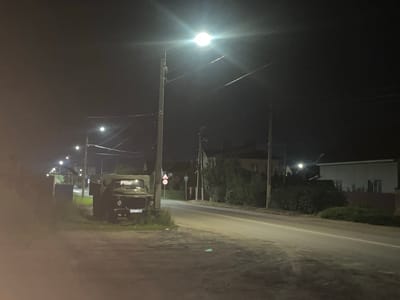 The image depicts a nighttime scene on a paved road in Borisoglebsk, Russia, illuminated by streetlights. In the foreground to the left, a military truck is parked on the unpaved shoulder, adjacent to a building structure and some vegetation. The truck is dark green, and its license plate, which reads "P631TK 36", is partially visible. Further down the road, houses and trees are visible on both sides. The road itself has a white line marking its center. Power lines run overhead, supported by utility poles. The overall atmosphere is quiet and still.