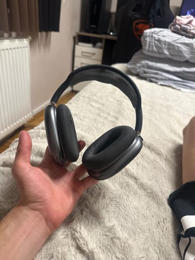 The scene is located in the city of Saraia, Bulgaria. The image presents an indoor setting, with a person's left hand prominently displaying a pair of Space Gray Apple AirPods Max headphones. The headphones feature a black mesh headband and black ear cushions, appearing to be in good condition. The hand and headphones rest upon a light-colored, textured, shaggy fabric surface.
In the background, a white panel radiator is visible on the left, adjacent to a light-colored wall. Above the radiator, brown and darker curtains are partially visible. To the right, a light-colored dresser or cabinet with two visible white drawers and silver pull handles is present, topped by a dark, rectangular object, possibly a monitor or television. Further right, a dark object with an orange, stylized cougar head logo is partially visible. A stack of folded textiles, including a pink garment, a striped gray item, and a plain gray item, is situated on what appears to be a bed. On the far right, a bare leg and a foot wearing a black and white sock or slipper are partially visible.