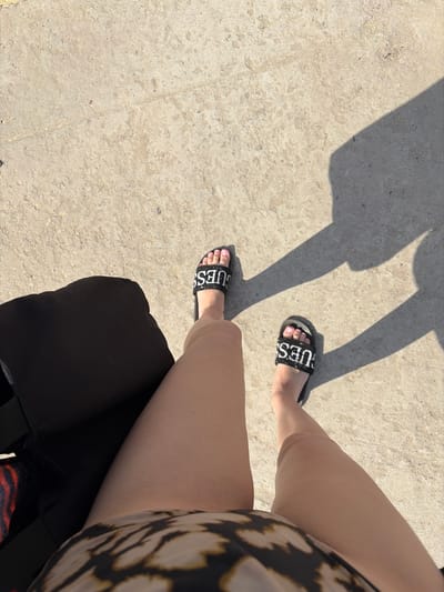 A pair of legs wearing black "GUESS" slide sandals are visible in the foreground, with the left leg closer and the right leg slightly further away. The sandals have white lettering on black straps and the toes are painted with light pink nail polish. The individual is wearing a patterned garment on their upper body, which is mostly out of frame. They are sitting or resting on a dark surface, possibly a stroller or bag, which takes up the lower right portion of the image. The background consists of a textured, light-colored concrete surface, suggesting an outdoor setting. Long shadows are cast on the concrete, indicating it is daytime, likely sunny. The lighting suggests it could be late morning or afternoon. The overall scene implies a relaxed moment, perhaps during a casual outing or while waiting. There are no explicit emotional cues or actions depicted beyond the stillness of resting. The visible text on the sandals is "GUESS". The location context is Novomykolaivka, Ukraine.