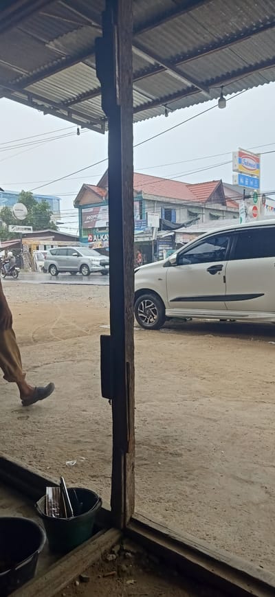 The image depicts an outdoor scene with a rustic wooden structure in the foreground, likely part of a shelter or building. Through the opening of this structure, a street scene in Uteungeulinggang, Indonesia, is visible. The sky is overcast, suggesting potentially rainy weather.

In the street, a silver SUV is parked, and a white SUV is driving past. A person is walking on the left side of the frame, with their lower legs and feet visible, wearing tan pants and dark grey slip-on shoes. Further down the street, a motorcycle with a rider is seen. Several buildings with tiled roofs line the street, some displaying signs, including one for "Indomaret" and another with "ALKA..." visible. The ground appears to be dirt or gravel.

Within the foreground shelter, a dark green bucket is prominently placed, containing what appears to be a metal object, possibly a pen or tool, and a piece of reflective material. To the left of this bucket, another dark-colored bucket is partially visible. The wooden structure is weathered, with visible grain and some damage. The overall impression is of a functional, perhaps somewhat informal, roadside environment.