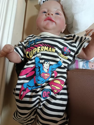 An overhead, medium shot shows a baby doll wearing a black and white striped onesie with a Superman graphic. The doll's head is tilted slightly to the right, with its blue eyes open and its mouth slightly agape. Its skin is pale, and its cheeks are flushed pink. The onesie features a large Superman logo on the chest and a smaller graphic of Superman in flight on the left side of the chest. The doll's arms are outstretched, and its right hand is visible, with painted blue fingernails. The doll is lying in what appears to be a cardboard box lined with white fabric. To the right of the doll, a brown, textured surface is visible, possibly a purse or a piece of luggage. The background is a light brown, and a white, crinkled fabric is visible behind the doll's head. The image is taken indoors, with neutral lighting. This scene has no relation to the city of Chinaz, Uzbekistan.