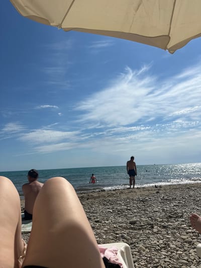 The image depicts a beach scene in Lermontovo, Russia, under a clear blue sky with scattered white clouds. In the foreground, parts of a person's legs are visible, suggesting they are lying on a beach chair. A beige umbrella shades the upper portion of the frame. The beach itself is covered in grey pebbles. In the middle ground, several people are visible. One man stands facing the sea, wearing dark shorts. Further out in the water, another person, wearing a red swimsuit, is wading. In the background, the calm sea stretches to the horizon, with the sun glinting off its surface. To the left, a person wearing a dark cap sits facing the water. The overall atmosphere is relaxed and summery.
