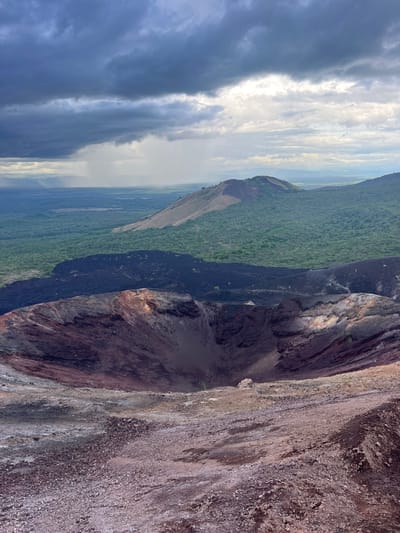 This is a vertical, landscape-oriented photograph taken from a high vantage point, likely the rim of a volcanic crater. The foreground is dominated by the steep, rocky slope of the volcano itself, showing layers of reddish-brown and grey volcanic rock, interspersed with gravel and smaller stones. There are a few sparse patches of green vegetation clinging to the rock face in places.

Beyond the immediate rim of the crater, the scene transitions to a lush, green forest covering the lower slopes of the volcano and extending into the distance. The forest appears dense and vibrant. Further in the background, the landscape flattens out into a more subdued, possibly cultivated or open terrain, under a wide, dramatic sky.

The sky is a prominent feature, with a mix of dark, ominous storm clouds on the left side and brighter, partly cloudy conditions on the right, suggesting a transition in weather or a dynamic atmospheric scene. Sunlight breaks through in some areas, illuminating the clouds and casting a soft light on the landscape.

The time of day appears to be late afternoon or early evening, given the quality of the light and the dramatic cloud formations. The overall impression is one of natural grandeur and the raw power of volcanic geology contrasted with the resilience of plant life. There is no text visible in the image. The location is Rota, Nicaragua, which is known for its volcanic features.
