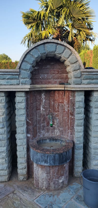 The image shows an outdoor water fountain structure with a large palm tree overhead. The fountain is constructed with stone and brickwork, featuring an arched niche at the top, a faucet, and a basin. To the right of the fountain, a dark grey bucket is visible on the stone patio. The background includes greenery and a clear blue sky. The scene is set in a garden or yard area.