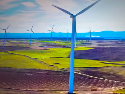 The image depicts a wind farm situated in a rural landscape, likely within the Basingstoke and Deane area of the United Kingdom. Numerous wind turbines, some closer and larger than others, stand tall in the open fields. The landscape alternates between bright green fields, possibly crops, and areas of brown, suggesting different land use or seasonal changes. In the background, a mountain range is visible under a pale blue sky with some cloud cover. There are no people visible in the scene. Roads and paths are subtly present in the foreground. The environment appears to be well-maintained and devoted to renewable energy production.