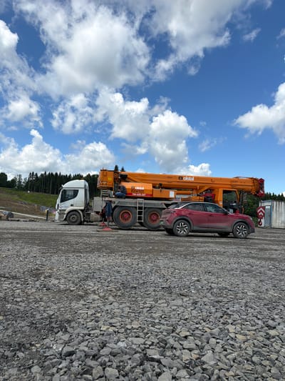 A red SUV is parked on a gravel lot in front of a large orange crane truck. The crane truck has "olstal" printed on its side, along with a phone number. A person is seated on the crane truck, appearing to be working. In the background, there is a forested hillside and a bright blue sky with scattered white clouds. A white semi-truck is also parked next to the crane. The scene appears to be a construction or industrial site.