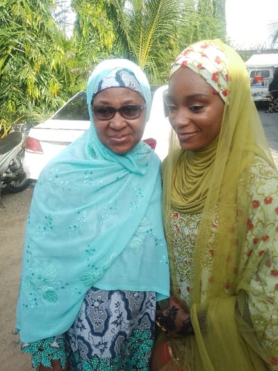Two women are positioned in the foreground of an outdoor scene in Unguwa Uku Kauyen Alu, Nigeria. The woman on the left is an older adult, wearing dark-framed sunglasses, a light blue head covering with an underlying blue and white patterned band, and a matching sheer light blue shawl adorned with light green floral embroidery and sequins. Her dress features a dark blue and white abstract floral pattern with a distinct green patterned hem. She is smiling and facing the camera directly. The woman on the right, a younger adult, is wearing an olive-green layered head covering (hijab) and a cream-colored long-sleeved dress extensively embroidered with gold, red, and green floral motifs. A sheer olive-green veil is draped over her head and shoulders. Her right hand, visible at waist level, displays intricate dark henna designs and gold bangles. She is looking downwards and slightly to her right. The background features dense green foliage, including palm fronds, behind the subjects. A white sedan is partially visible behind the left woman, with a silver and black motorcycle partially visible to its left. A white tent or canopy is visible in the upper right background. The ground is unpaved, light brown earth. The scene is illuminated by bright natural daylight.