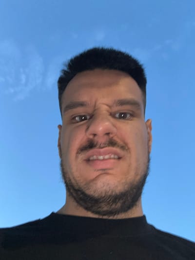 The image is a close-up, low-angle selfie of a man with dark hair, a mustache, and a beard. He is wearing a black t-shirt and making a grimacing facial expression with his teeth bared. The background is a clear blue sky with a few wispy clouds, suggesting an outdoor setting. The lighting indicates it is daytime, possibly late afternoon or early evening. The man's features are prominent due to the close-up nature of the photograph. No specific location in Roquetas de Mar, Spain, can be identified from the image itself.