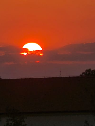 The media file depicts a dramatic sunset over Holíč, Slovakia. The sun, a bright orb with an intense orange glow, is partially obscured by dark, textured clouds. The sky transitions from a vibrant orange near the sun to a muted purple and gray as it moves away.
To the right of the sky, a dark, indistinct mass suggests a landscape, possibly trees or buildings, rendered almost silhouetted against the twilight. A thin, vertical structure, resembling a fence or utility pole, is visible in this darker area, adding a man-made element to the natural scene. The overall mood is one of natural beauty and the transition from day to night. The time of day is clearly sunset. There is no visible text in the image.