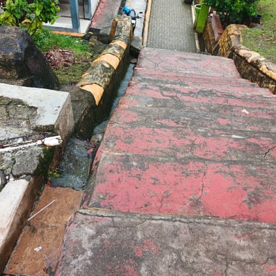 The image shows a set of concrete stairs painted a faded red color. The stairs are located in Pameungpeuk, Indonesia, at the coordinates -7.637 latitude and 107.731 longitude. The stairs are cracked and worn, and there is a small stream of water running down the side of the stairs. The stairs are surrounded by concrete and brick walls, and there is a small amount of vegetation growing in the area. The image was taken from a high vantage point, looking down at the stairs.  The photo is taken from a high angle, looking down on the stairs. The stairs are in a worn state, and there is a small stream of water flowing down the side.  The stairs are surrounded by concrete and brick walls, and there is a small amount of vegetation growing in the area.  There is no indication of anyone present at this scene.