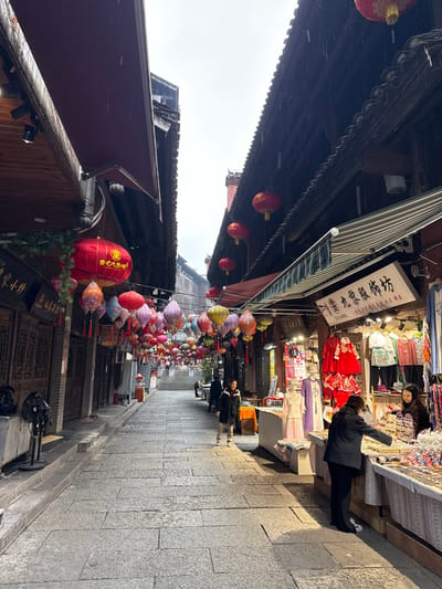 The image displays a narrow, stone-paved commercial street in Pengshui Miao and Tujia Autonomous County, China, under an overcast sky with visible light rain. The street gently inclines uphill, flanked by traditional multi-story buildings characterized by dark wooden facades and tiled roofs. A dense canopy of colorful lanterns, including round red designs and ornate pleated variations, is strung across the street overhead.
On the right side, an open-front stall, identified by a white sign bearing Chinese characters including "九娘服饰坊" (Jiuniang Clothing Workshop), showcases garments such as red traditional dresses and various patterned apparel. Two women are present at this stall: one, wearing a black jacket and pants, examines items on a display table, while another woman, likely a vendor, stands behind the table. Additional commercial stalls are visible further along the street on the right.
The left side of the street is lined with buildings featuring dark wooden exteriors and closed doors. A black floor fan is positioned on the sidewalk in the foreground. In the mid-ground, a man dressed in a dark jacket walks uphill on the stone pavement. Another individual is seated on a structure further along the left side of the street. The stone pavement appears damp.