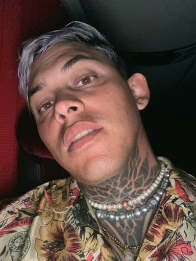 A person with short, gray-tipped hair lies at an angle, smiling slightly. They have brown eyes and a noticeable geometric tattoo covering much of their neck. Several strands of pearls and silver beads adorn their neck. They are wearing a patterned shirt with floral and text-based designs. A dark surface and a red textured material are visible in the background.