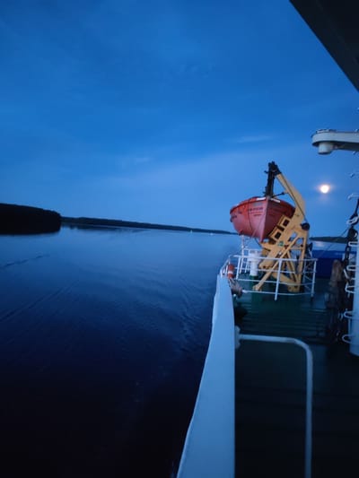 The image captures a twilight scene from the deck of a vessel at sea, likely near Matveevskoe, Russia. The sky is a deep, clear blue, transitioning to a darker shade at the horizon. A crescent moon is visible in the upper right quadrant of the sky. On the right side of the frame, a portion of the ship's deck is visible, featuring a bright orange lifeboat suspended from a yellow davit. The lifeboat appears to be intact and secured. The water is calm and dark blue, reflecting the twilight sky. To the left, a dark, tree-lined shoreline stretches across the horizon, indicating landmass nearby. The overall impression is one of quiet navigation as day transitions to night. There are no people visible in the image.