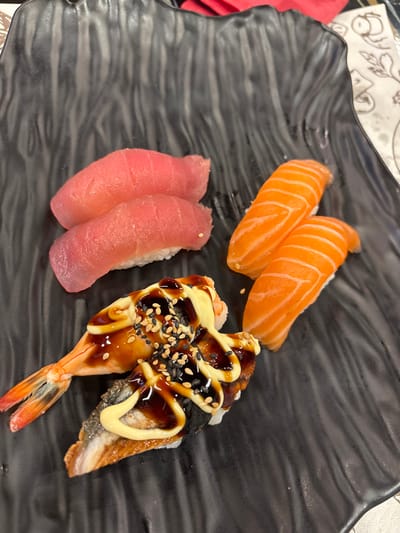 A close-up, slightly elevated perspective depicts six pieces of nigiri sushi arranged on a black, undulating-surface platter, situated within a dining establishment in Plasencia, Spain. The upper left quadrant of the platter displays two tuna nigiri, characterized by their uniform pink-red coloration atop white rice mounds. To their right, in the upper right quadrant, two salmon nigiri are visible, distinguished by their bright orange hue and distinct white striations, also presented on white rice. The lower left quadrant features two specialty nigiri: one comprises a cooked shrimp, adorned with a dark, viscous sauce, a lighter, opaque sauce, and an application of black and white sesame seeds over the shrimp and rice; the adjacent piece, an eel nigiri, is similarly prepared with dark and light sauces, and sesame seeds. A partial view of a white patterned surface is visible in the top right background, indicating a table covering.
