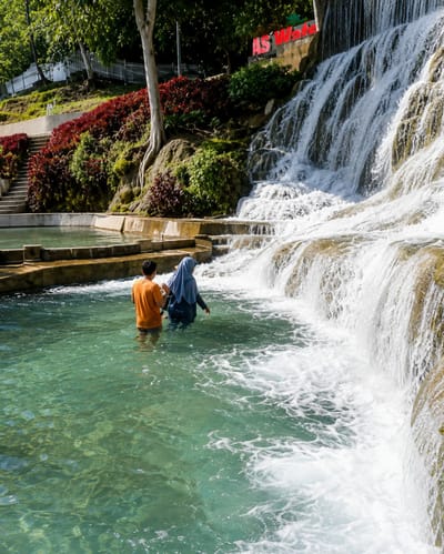 In the city of Silaiyajae, Indonesia, a scenic shot captures two individuals standing in the shallow, turquoise waters of a tiered waterfall. The person on the left, wearing an orange t-shirt, faces away from the camera, while the person on the right, adorned in a blue hijab and dark clothing, also has their back to the viewer. The waterfall cascades down a series of rocky ledges, creating a foamy, white expanse of water. Lush greenery and red-leafed plants adorn the sloped terrain surrounding the waterfall, with a prominent tree and visible roots anchoring the earthy tones. In the background, a white fence and stairs suggest a developed recreational area, and red lettering on a stone structure at the top of the waterfall reads "AS WATA". The water is clear enough to reveal the rocky bottom, and ripples on the surface indicate gentle movement. The scene is bathed in natural sunlight, casting subtle shadows and highlighting the vibrant colors of the landscape.