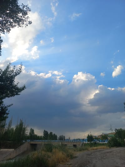 The media file depicts a scene in Kasansay, Uzbekistan. The sky is the dominant subject, with a mix of bright blue areas and dramatic, dark storm clouds. Sunbeams pierce through some of the lighter clouds, creating a dynamic contrast. The weather appears to be changing, with the possibility of rain or a storm approaching.
In the foreground, the tops of lush green trees frame the lower left and right edges of the image. The trees suggest it is springtime or summer.
The background reveals more of the setting. A road runs along the right side, appearing to be unpaved and dry, with dry grasses and vegetation alongside it. Further into the background, there's a glimpse of buildings and structures, possibly a residential area or a small town. A fence or railing with blue accents is visible, suggesting a pathway or boundary. There are also indications of more trees, forming a line along the horizon. The overall impression is a rural or semi-urban environment.
The time of day is likely late afternoon or early evening, given the angle of the sunbeams and the interplay of light and shadow on the clouds. There are no people or specific activities clearly depicted. The scene evokes a sense of anticipation due to the impending weather. No visible text is present in the media file.