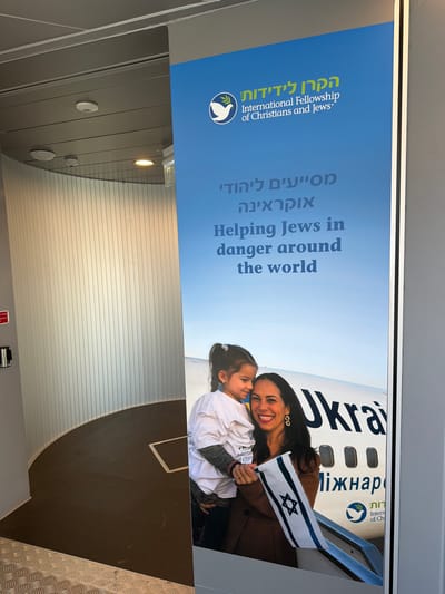 The image shows a large blue banner with text in Hebrew and English, and a photograph of a woman and a child. The banner is vertical and appears to be displayed indoors, possibly in a public building or airport terminal. The text at the top of the banner reads "הקרן לידידות" and "International Fellowship of Christians and Jews". Below that, in Hebrew and English, it says "מסייעים ליהודי אוקראינה" and "Helping Jews in danger around the world". The photograph features a woman smiling and holding an Israeli flag, with a young girl nestled close to her. Behind them is the tail of an airplane with "Ukrain" visible on it. The environment includes a curved wall with vertical paneling and a section of dark flooring. There are also some ceiling fixtures visible.