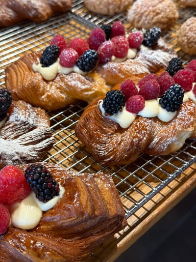 A close-up, eye-level shot captures several raspberry and blackberry danishes arranged on a metal cooling rack, which sits on a wooden surface. The danishes, which appear to be freshly baked, are golden brown and flaky with a dusting of powdered sugar. Each pastry is topped with a dollop of white cream and adorned with fresh raspberries and blackberries. In the background, out of focus, are more pastries, some of which appear to be similar danishes or croissants. The lighting suggests an indoor setting, possibly a bakery or cafe. The scene is entirely focused on the pastries, highlighting their texture and fresh fruit toppings. No people are visible in the image. The location is not identifiable from the image itself, therefore its presence in Kfar Monash, Israel, cannot be confirmed.