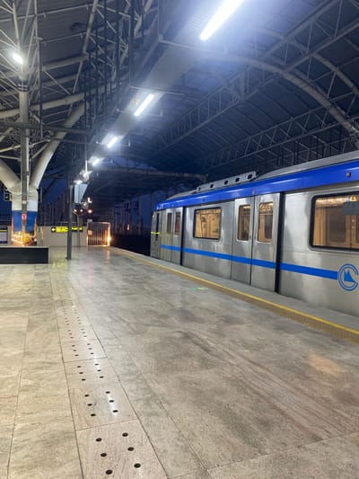 An outdoor metro train station platform at St. Thomas Mount-Pallavaram Cantonment Board, India. The platform is made of light-colored stone tiles with a series of circular indentations forming a line along the edge. A silver and blue metro train is stopped at the platform, with its doors closed. The train has illuminated windows, and a blue stripe runs along its length. Above the platform, a large, industrial-style roof structure with metal beams and corrugated metal panels is visible, along with fluorescent lighting fixtures. The sky in the background appears to be twilight or early evening. Signs with yellow arrows and text are mounted on poles, and there is a small circular mirror reflecting light on one of the poles. The overall scene is well-lit by artificial lights, creating a sense of a functional, modern transit hub.