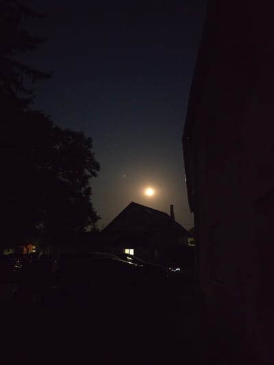 The image depicts a nighttime scene in Kostelec na Hané, Czechia. A bright full moon is visible in the dark sky, positioned above the silhouette of a house. The house has a single lit window, suggesting occupancy. To the left of the house, a large, dark tree is silhouetted against the sky. In the foreground, the dark shapes of several parked cars are discernible, with some of their lights faintly glowing. A portion of a building is prominent on the right side of the frame, also in silhouette, obscuring the view to its right. The overall atmosphere is dark and quiet, illuminated by the moon and the single interior light.