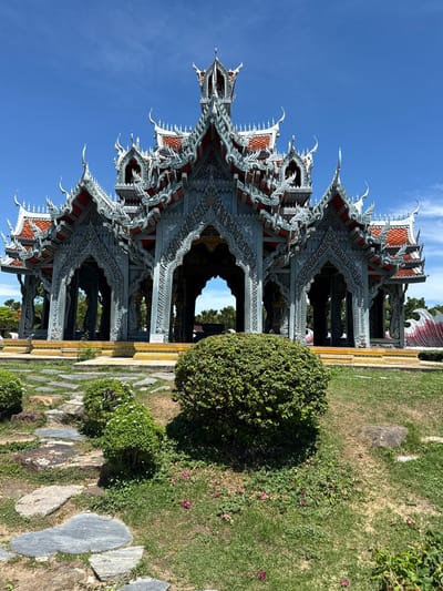 The image shows an ornate, multi-tiered pavilion structure with a gray facade and red roof accents. It is surrounded by meticulously maintained green lawns and stone pathways. The location is Bang Pu Subdistrict Municipality, Thailand. A rounded, neatly trimmed bush is positioned to the right of the structure. The sky is clear and blue.