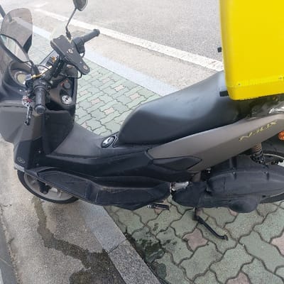 A dark gray and black Yamaha NMAX maxi-scooter is parked on a patterned greenish-gray paver sidewalk next to a concrete curb. The scooter features a clear windshield, handlebars with a mounted phone holder, and a key ring with multiple keys and charms. A prominent, bright yellow rectangular delivery box is affixed to the rear of the scooter, behind the black seat. The scooter's side stand is extended, supporting the vehicle. In the background, an asphalt road with a white painted line is visible. The scene is outdoors, in 김포시, South Korea. No individuals or active interactions are present.