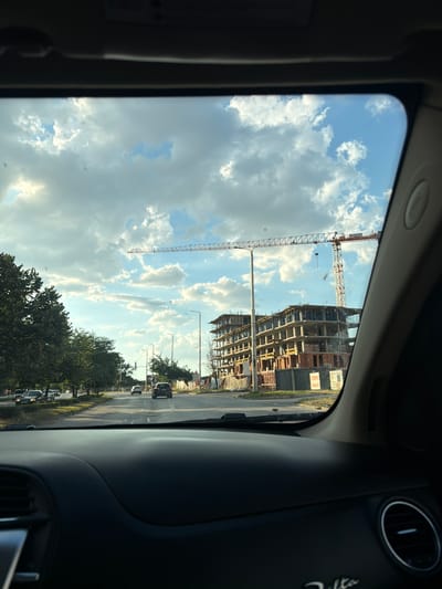 This image captures a view from inside a vehicle looking out at a street scene in Georgi-Dobrevo, Bulgaria. The perspective is that of a passenger or driver, with the dashboard and windshield frame visible in the foreground.
The outdoor scene is dominated by a bright, partly cloudy sky with dramatic cumulus clouds. A long construction crane, typical of urban development, stretches horizontally across the middle ground. To the right, a multi-story building under construction is visible, showing concrete framing and some brickwork.
The street itself is paved and appears to be a main thoroughfare with multiple lanes. Several cars are driving on the road, including a dark SUV in the right lane and other vehicles further down. Trees line the left side of the street, providing a green contrast to the construction. Streetlights and traffic signals are also present.
The overall impression is one of an active urban area undergoing development, under a dynamic sky.