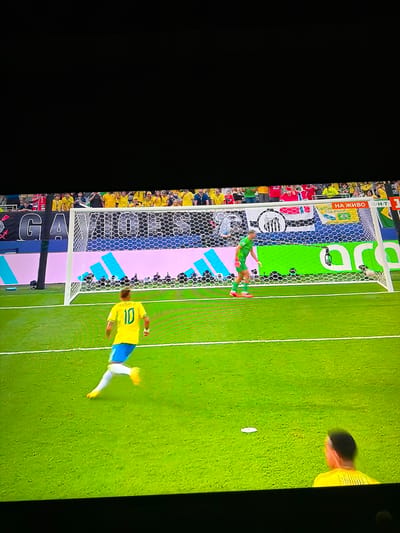 This image captures a football match, likely a penalty kick, on a bright green field. A player wearing a yellow jersey with the number 10 and blue shorts is in motion, with his right leg extended as if striking a ball, which is not clearly visible but implied by his posture. He is facing towards a goal defended by a goalkeeper in a green jersey and orange socks. The goalkeeper is positioned near the goal line, facing the penalty taker. Behind the goal, spectators are visible in the stands, many wearing yellow. A banner with text and logos is draped across the stands. The environment appears to be a stadium setting with floodlights illuminating the field. This scene is not located in the city of Ognianovo, Bulgaria; there are no visual cues in the image to suggest this location.