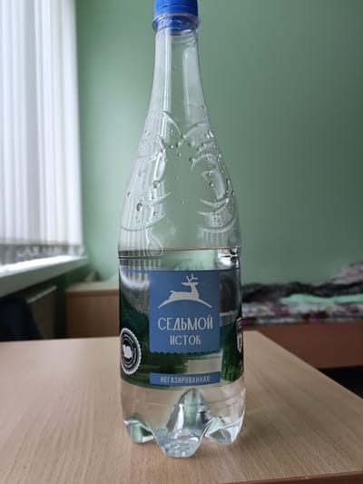 A clear plastic bottle of water, with a blue cap, sits on a wooden table. The label on the bottle is blue and white, featuring a white deer silhouette and the text "СЕДЬМОЙ ИСТОК" which translates to "Seventh Spring". Below this, the word "НЕГАЗИРОВАННАЯ" indicating "still" water. The bottle appears to have condensation on its exterior. The background shows a green wall and a window with white blinds. The image is a still life, with no people or actions depicted. The specific location in the city of Ключи, Russia, is not ascertainable from the visual information provided in the image.