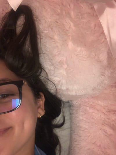 A person with long, dark hair lies on a soft, pink, furry surface. The person is wearing glasses with black frames. The left side of their face, including an eye, cheek, and part of their mouth, is visible. Several rings adorn their ear. The background features a blend of light pink and white textures.