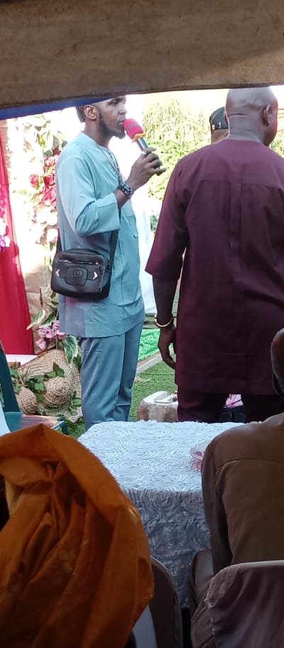 The image depicts an outdoor gathering in Udi, Nigeria. A male individual, positioned left of center, wears a light blue tunic and matching trousers, along with multiple dark beaded bracelets on his left wrist and a dark shoulder bag with a front decorative element. He holds a red and silver microphone to his mouth, facing right in profile. To his immediate right, a bald male wears a dark maroon tunic and a gold-colored bangle on his right wrist, with his back partially to the viewer. In the lower right foreground, a third individual is partially visible, seated, with an orange fabric draped over their person. The foreground includes a table covered with a white, textured tablecloth. The background features event decorations: red fabric drapes on the far left, artificial foliage with pink flowers, and woven straw-like objects, one large and round with green leaves. A green mat-like surface is on the ground, with an unidentifiable light-colored decorative object further back. Natural green foliage is visible in the distant background, and a wooden beam stretches across the top of the frame.