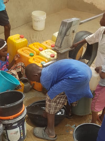 People are collecting water in what appears to be an urban environment in Dutsinma A, Nigeria. Individuals fill various containers, including plastic jerrycans (predominantly yellow) and buckets, using a pump. The area around the pump is muddy, with several containers scattered nearby. One person is actively filling a black bucket while another appears to be operating the pump. A tire is present near the pump mechanism. The walls of a building form part of the backdrop.