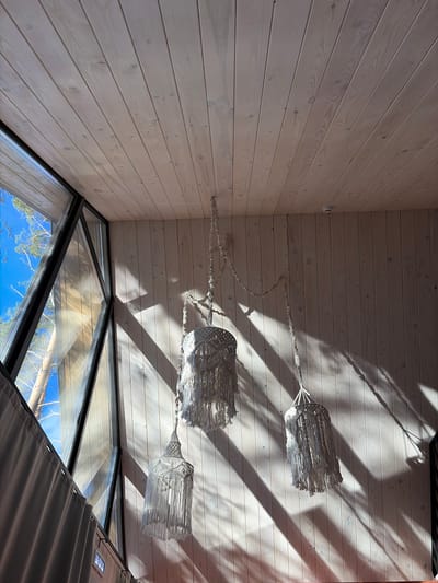 Captured from a low-angle perspective, the photograph showcases the interior of a structure in Мишкино, Russia. The walls and ceiling are clad in light-colored wood paneling. Natural light, filtered and patterned by external elements, casts distinct shadows across the ceiling. A multi-tiered macramé light fixture hangs centrally. Below, angular windows frame a view of the clear, blue sky and treetops. A section of neutral-toned curtains is visible on the right edge of the frame.