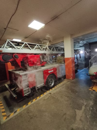 A red fire truck with a ladder, labeled "MAGIRUS," is parked indoors in what appears to be a garage or fire station. Wisps of white steam or smoke are rising from the truck's body. The environment includes a concrete floor with yellow and black hazard stripes marking the area around the truck. Overhead, fluorescent lights illuminate the space, and electrical conduits are visible on the ceiling. In the background, another vehicle and garage doors are partially visible. The scene suggests a potential test, cleaning, or minor malfunction of the fire truck, as indicated by the steam. The location is identified as Tukums, Latvia.