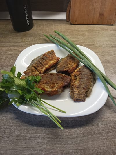 A close-up, overhead shot displays a white plate on a wooden surface. The plate holds four pieces of fried fish, a bunch of parsley, and several green onions. In the background, a black thermal mug with "WOODY PEOPLE" printed in white is visible, as well as a wooden cutting board. The scene is consistent with a home kitchen setting in Volgodonsk, Russia.