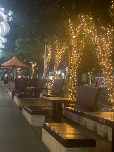 This image depicts an outdoor seating area at night, likely in an urban setting. The area is lined with trees adorned with numerous small, warm-toned string lights, creating a festive and inviting ambiance. In the foreground, several wooden picnic tables with attached benches are visible. These tables and benches are constructed with dark wooden seating and tabletops, supported by concrete bases. The pathway in front of the seating area appears to be paved. In the background, a large, modern, white, curved structure can be seen, possibly a part of a building's architecture or a sculptural element. Further back, under an orange awning, patrons are seated, suggesting a restaurant or cafe setting. The overall scene is illuminated by the string lights and ambient artificial light.
This scene is located in the South Bank precinct of Brisbane, Australia, specifically at The Jetty South Bank, known for its riverside dining and entertainment options. The characteristic modern architecture and the presence of outdoor dining facilities are indicative of this popular Brisbane location.