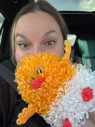 The image is a selfie taken inside a vehicle. The focal point is a woman's face, partially obscured by a handcrafted, brightly colored plush toy. The toy appears to have an orange, curly texture with red and white accents. The woman has brown eyes and light brown hair with blonde highlights. She is wearing a seatbelt. Behind her, blurry details suggest a vehicle interior with additional items, possibly green bags. The location, Татарские Челны, Russia, cannot be confirmed solely from visual cues in the image.