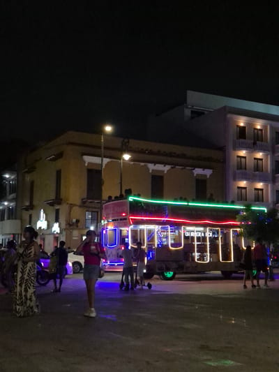The image depicts a street scene at night in Veracruz City, Mexico. The street is illuminated by neon lights and street lamps. In the background, there is a building with multiple stories and lit windows, as well as a building with a neon sign that says "CHURRERIA FOR..." (the rest of the word is not visible). People are walking and standing on the sidewalk. Some are riding scooters or bicycles. A woman is walking on the street. A variety of businesses or shops appear to be open, indicated by lights and signage. The overall atmosphere is lively with the presence of people and bright lights.
