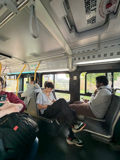 This vertical image captures the interior of a public transit bus during the daytime, likely in Longueuil, Canada, as passengers go about their commute.

In the foreground, a person's hands are visible holding a green smartphone, positioned next to a black "Samsonite" backpack. Further into the bus on the right side, a person with dark, curly hair, wearing a gray hoodie and brown pants, is seated by a window, looking down at their phone while wearing white earbuds. Their feet, clad in distinctive black and light blue sneakers with white soles, are visible on the bus floor. Directly across the aisle, another person with curly hair, dressed in a light gray t-shirt and wearing a seatbelt, is also seated by a window, appearing relaxed or looking out. The bus interior features gray and white panels, silver support poles, and black textured seating.

Outside the windows, daylight illuminates trees, buildings, and a glimpse of an urban or suburban landscape.

Key textual details reinforce the setting: an overhead sign clearly reads "SORTIE D'URGENCE / EMERGENCY EXIT," with partial instructions "THEN PUSH KN..." and "PRESSER LE BOUTON POUR OUVRIR" in both English and French. Further forward, signs display "PAIEMENT COMPTANT 1 PASSAGE BUS SA" (Cash payment 1 bus ride) and "CORRESPONDANCE" (Transfer), indicating payment and transfer information. A "NO SMOKING" symbol is also visible on a window. The overall atmosphere is typical of daily public transport.