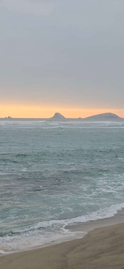 The image depicts a serene coastal scene in Punta Hermosa, Peru, under an overcast sky with a hint of orange peeking through near the horizon. The ocean is moderately choppy, with white-capped waves rolling towards a sandy beach. In the distance, several rocky islands rise from the sea. The foreground is dominated by the beach, with gentle waves lapping at its edge. The overall impression is one of tranquility and natural beauty.