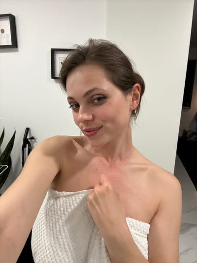 A woman is posing in a bathroom, partially covered in a white towel. She has brown hair pulled back and is looking at the camera. The bathroom has white walls with framed pictures and a plant visible in the background. The location in Kranj, Slovenia cannot be determined.