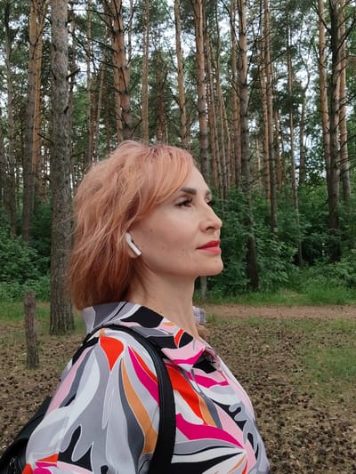 A medium shot captures a woman with salmon-colored hair in a profile view, facing right. She is wearing white wireless earbuds in her left ear and a black, white, pink, and orange patterned blouse. She has makeup on, including eyeshadow and lipstick. A black strap, possibly from a backpack, is visible across her shoulder. The background consists of a dense forest with tall, slender pine trees and lush green foliage. The ground is covered with dry pine needles and a dirt path is visible. The lighting suggests an overcast day. The image was taken in a park in the city of Chaikovsky, Russia.