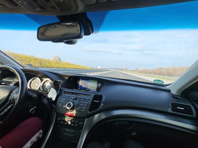 The image depicts the interior perspective from the driver's seat of a passenger vehicle traveling on a multi-lane asphalt highway in the vicinity of Malko Tranovo, Bulgaria. The car's dashboard, primarily black with grey accents, occupies the mid-ground. A black, multi-function steering wheel is visible on the left, partially obscuring the driver's left leg, which is clad in dark red leggings with a white patterned band. A black smartphone is vertically mounted on the central air vent. Above the climate and audio controls, a navigation screen displays a map with route data, showing "A1 Orizovo" on the interface. A red hazard light button on the control panel is illuminated. A silver key ring with keys hangs from the ignition area. The windshield, displaying streaks and spots, features a blue sun-shade tint at the top and a rearview mirror mounted centrally. Outside the vehicle, the highway extends straight into the distance, bordered by white guardrails and grassy embankments covered in yellowish-green foliage. Distant white advertising billboards and scattered white-blossomed trees are visible on either side of the road. The sky is light blue with sparse white clouds. Other vehicles are present as distant traffic on the highway. A passenger's legs are partially visible in the lower right foreground.
