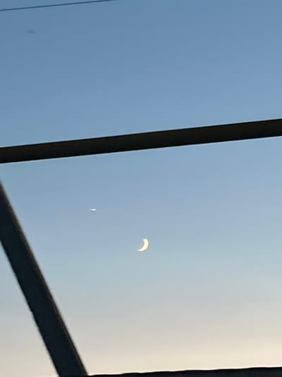 The image captures a partial solar eclipse seen from Sibay, Russia. The sky is a clear, pale blue, suggesting it is daytime, likely mid-morning or afternoon, with bright sunlight. A thin, crescent-shaped sliver of the sun is visible, indicating that the moon is partially obscuring it. There is also a faint, white, vertical line or streak in the sky below the crescent, its nature unclear from the image alone. The scene is framed by dark, thick, possibly metallic or wooden beams, suggesting the view is through a window or structure, perhaps a rooftop or an industrial setting. The overall impression is one of observation and a celestial event in progress. There is no visible text in the image.