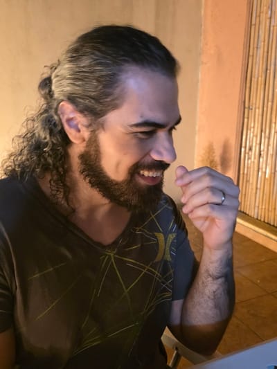 An adult male is depicted in a close-up, waist-up portrait taken in Villa San Andrés, Argentina. He is positioned facing right in profile, smiling with teeth visible and a dimple on his left cheek. He possesses long, dark, wavy hair pulled back into a small bun or ponytail, with loose curls framing his neck and ears, and a full, dark beard and mustache. He is wearing a dark grey V-neck t-shirt with a pattern of thin, light-colored geometric lines and a partial light-colored logo on the right chest, appearing to be a curved 'Hurley' emblem. His left hand is raised towards his mouth, with fingers slightly curled, and a plain silver-colored ring is visible on his left ring finger. His left forearm, covered in dark hair, rests on an unseen surface. The background features a plain, light tan wall to the left and a vertical screen or decorative element composed of light-colored bamboo rods to the right. A white surface is partially visible in the bottom right corner, likely a table. The scene is illuminated by warm, artificial light.